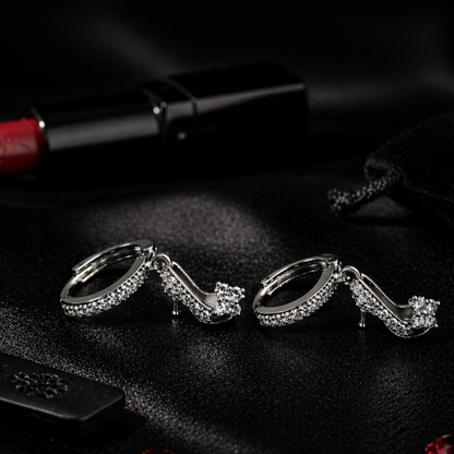 Silver high-heeled shoe earrings on a dark surface with red lipstick and gemstones.
