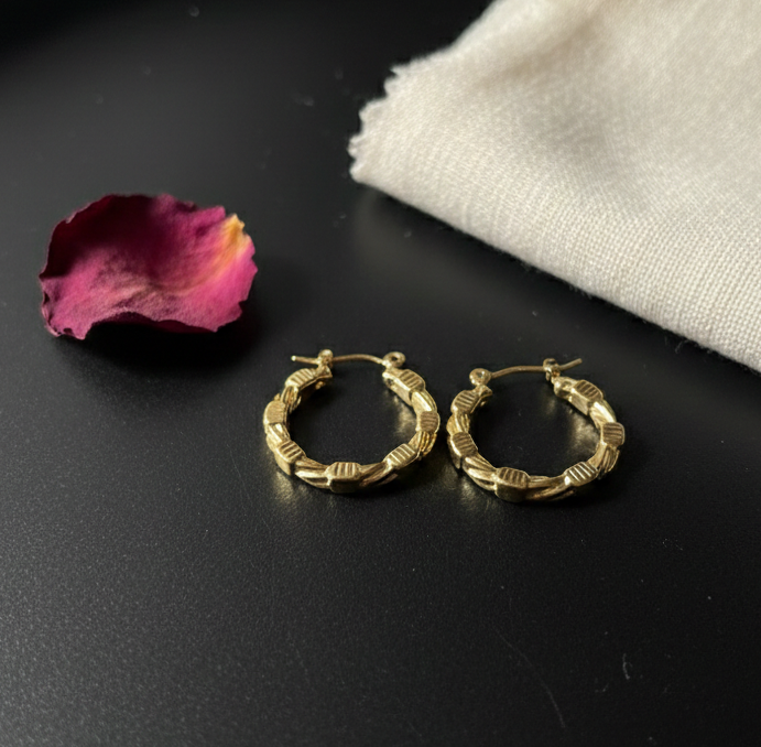 Gold hoop earrings on a black surface with a pink flower petal and white cloth.