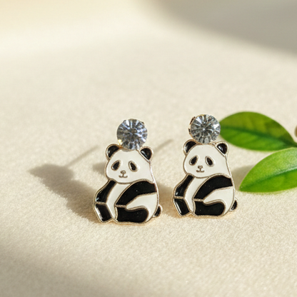 Panda-shaped earrings on a beige surface with a bamboo branch and leaves.